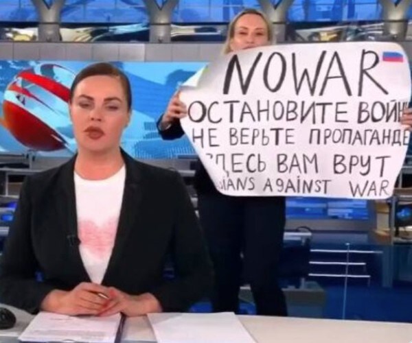 tv anchor with protestor behind her with sign reading no war in english with russian writing beneath it