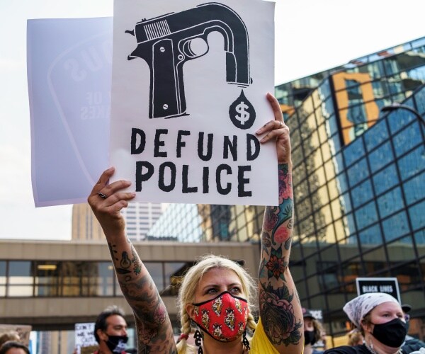 a woman in a mask holds a defund the police sign