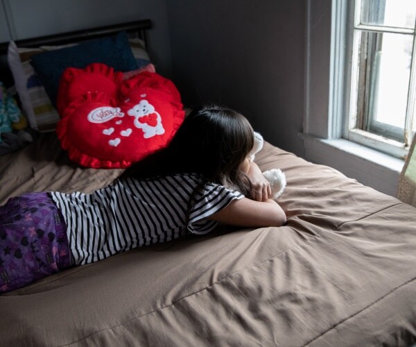 little girl hugs a stuffed animal and looks out the window of her bedroom