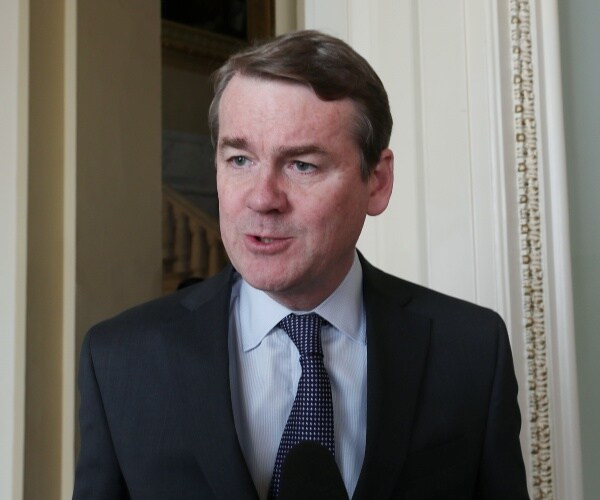 bennet in a suit and light blue shirt speaking into a mic