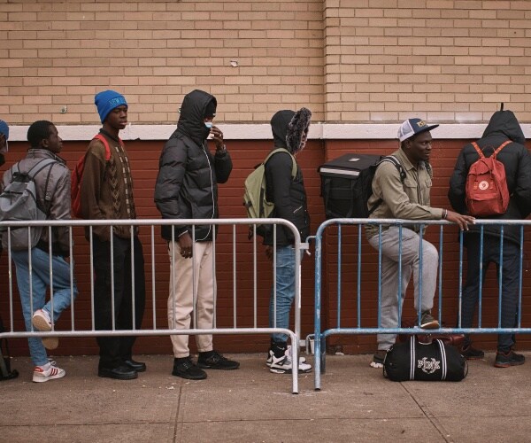 migrants in winter clothing stand on line behind gate