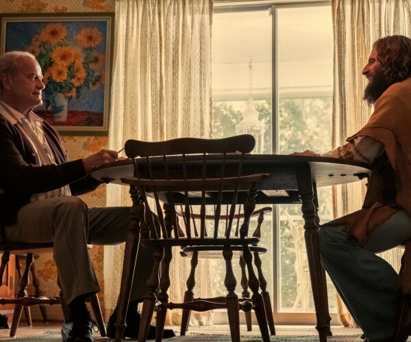 middle aged man in casual dress clothes sits at kitchen table with young man with long hair beard and hippie clothes