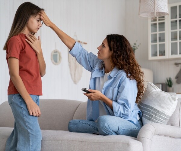 mother holding smartphone with one hand and other hand feeling sick daughter's forehead
