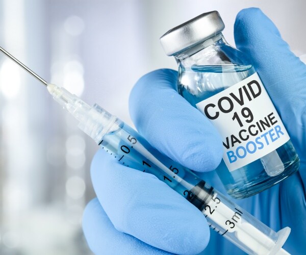 a hand with blue glove holding a vial labeled COVID-19 booster shot and a syringe