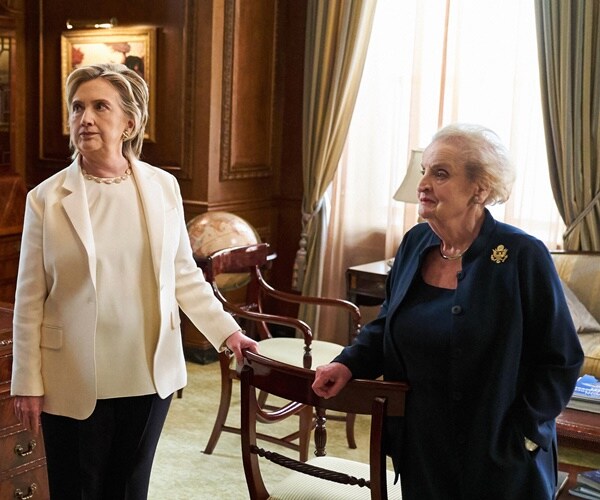 hillary clinton and madeleine albright listen to someone talking on a tv set.