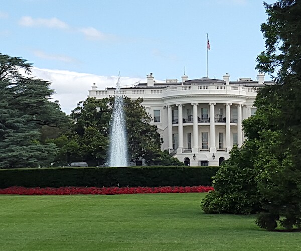 the white house is nestled between trees and behind a water fountain
