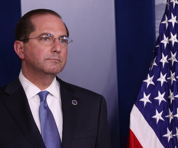 azar in a suit and blue tie standing near an american flag