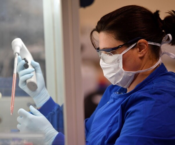 woman in a white face mask, goggles and blue uniform holds a tube wearing gloves