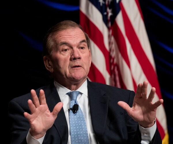 ridge in a suit and light blue tie sitting in front of a us flag