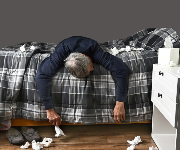 sick man laying on stomach sideways across bed, tissues all around on ground