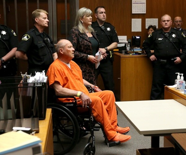 golden state killer in an orange outfit sits in a wheelchair with police surrounding him