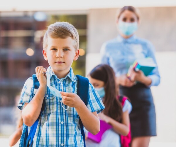 boy at school taking off mask, teacher in background with mask on