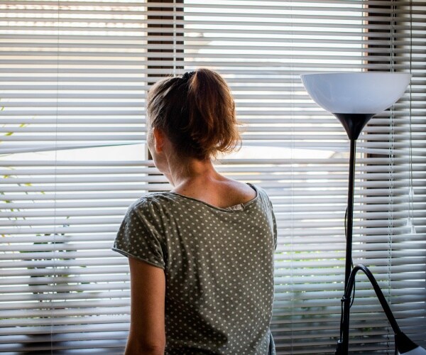 woman home alone isolating, looking outside through a crack in her blinds