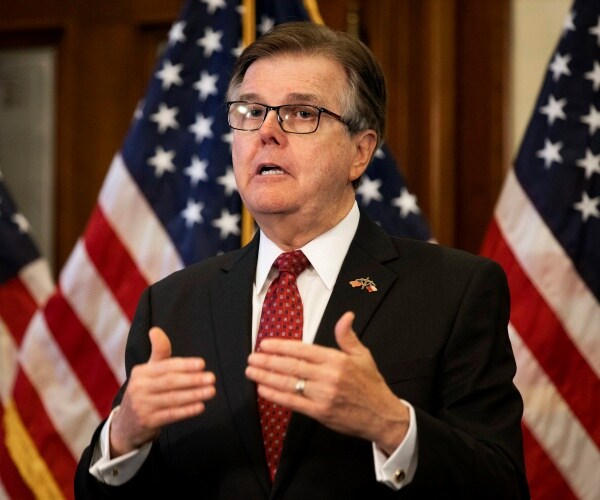 patrick in a black suit and red patterned tie standing in front of american flags