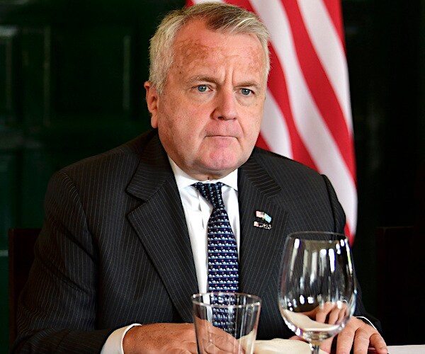 John Sullivan sits at a state dinner with an american flag in the background