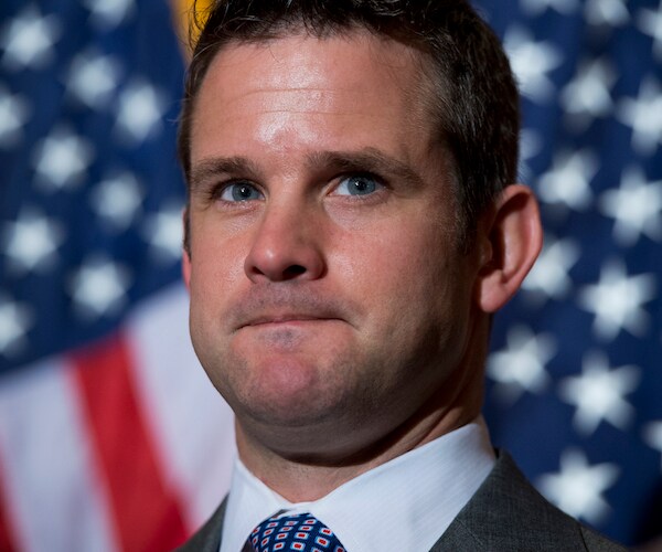 adam kinzinger bites his bottom lip while standing silent at the podium with an american flag in the background