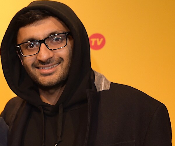 nimesh patel smiles, wearing glasses and a brown hoodie, which is draped over his head