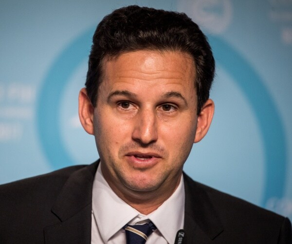 brian schatz in a black suit and striped tie speaking in front of a blue background