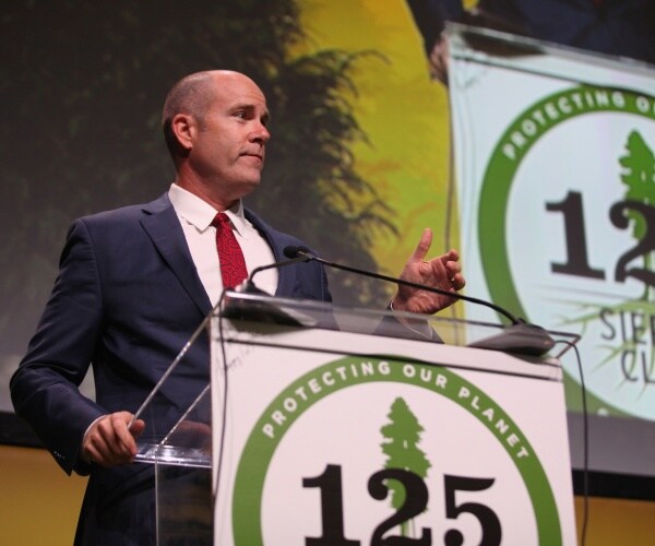 brune in a suit speaking on stage at a clear podium 