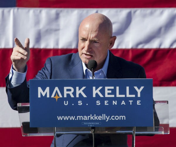 mark kelly is shown speaking from a podium that bears his campaign sign with the us flag in the background