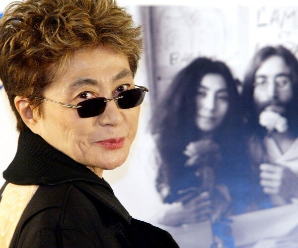yoko ono in a black top and small sunglasses standing near a picture of her and john lennon