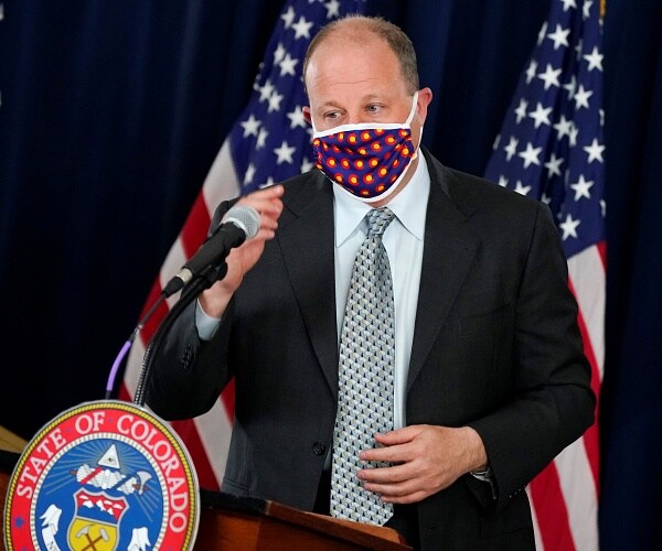 jared polis stands behind podium wearing mask