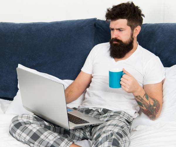 man lying in bed working on laptop computer with a cup of coffee in one hand