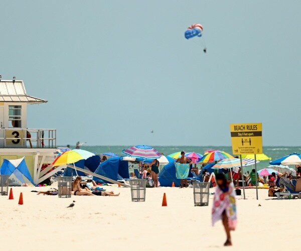 people sit under colorful umbrellas on the beach with a sign telling people to socially distance 