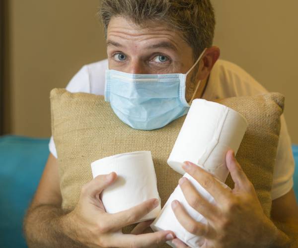 a man looking scared wearing a medical mask and clutching a pillow and rolls of toilet paper