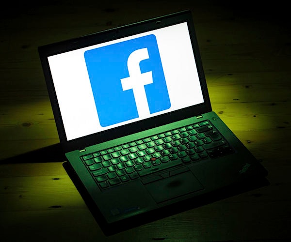 a laptop lays on a brown table in a dark room with the facebook logo on the screen