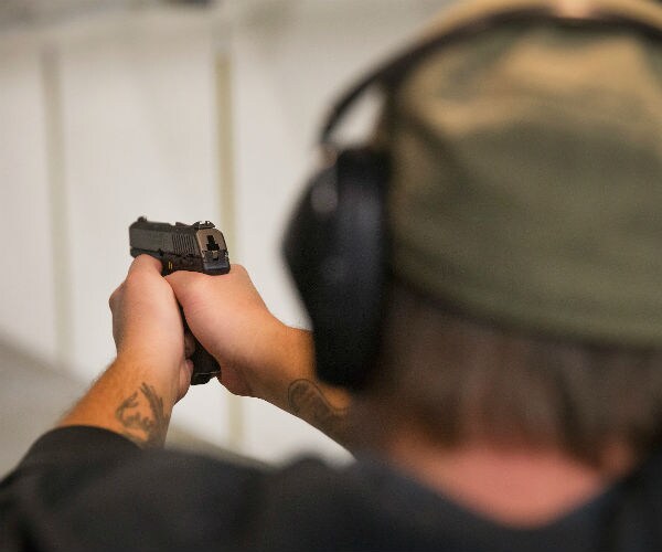a man wearing noise canceling ear muffs and a green hat is pictured from behind pointing a gun at a shooting range