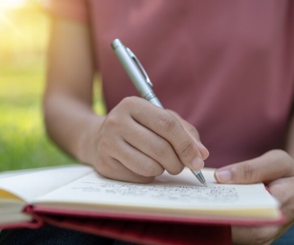 woman writing in a notebook with a pen wearing a pink shirt