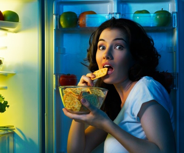 woman holding bowl of crackers, looking in refrigerator in dark kitchen