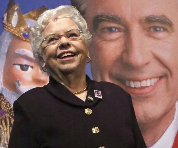 joanne rogers standing in front a poster of her husband and one of his puppets king friday the thirteenth