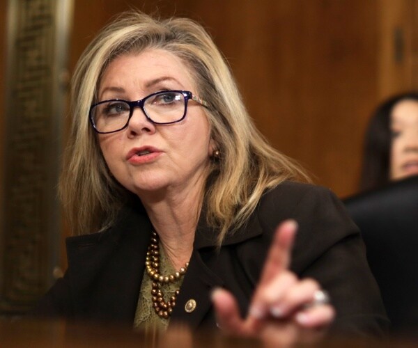 blackburn in a black coat and green top wearing glasses