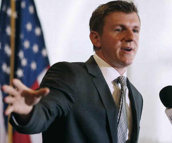 james o'keefe extends the five fingers of his right hand while speaking to reporters with an american flag behind him