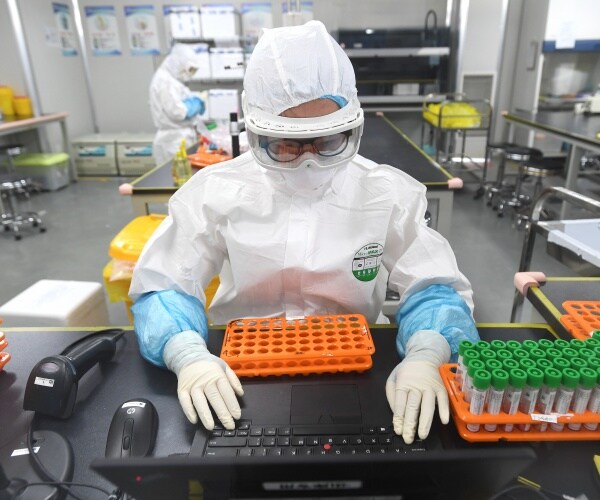 researcher in white coat and mask at a lab in china types on a laptop with many small test tubes next to him