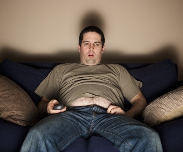 an overweight man on couch watching television with bored look on face