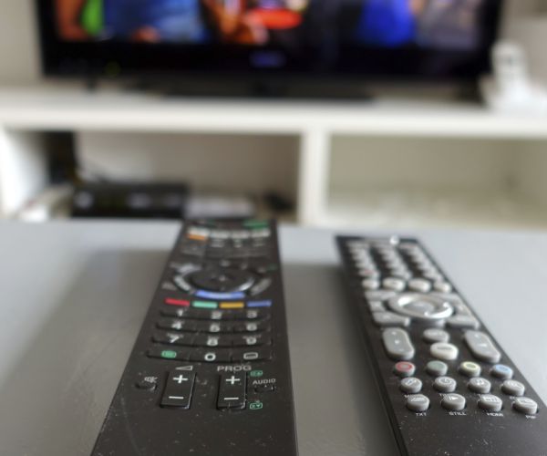 two television remote controls sit on a white table with a television on in the background