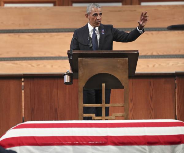 obama raises his arm while speaking with a flag draped casked below him