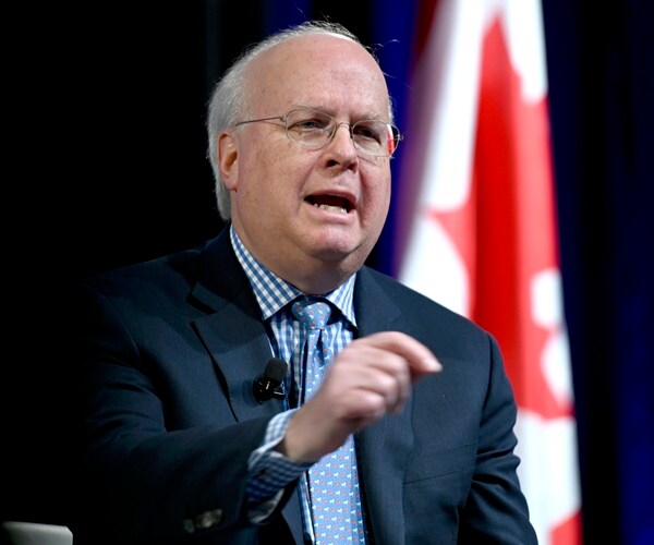 karl rove sits on a stage and speaks to an audience in front of a canadian flag.