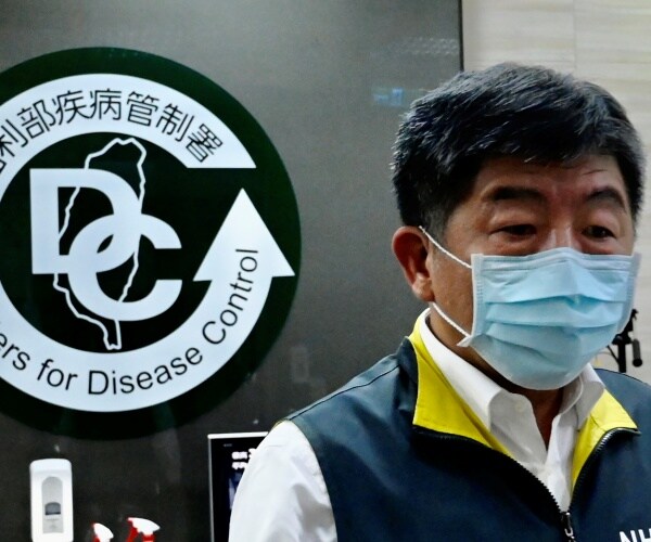 health minister in a gray and yellow jacket and white shirt walks by taiwan cdc logo