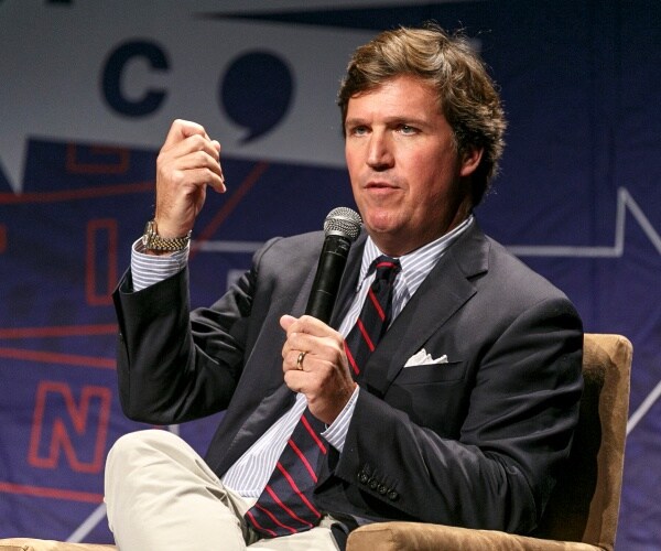 carlson in a black suit and navy blue and red striped tie speaking at politicon