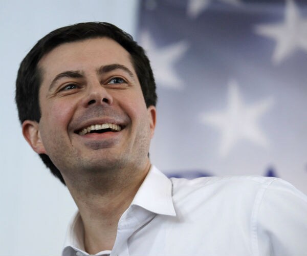 pete buttigieg looks to his left and smiles wide in front of an american flag