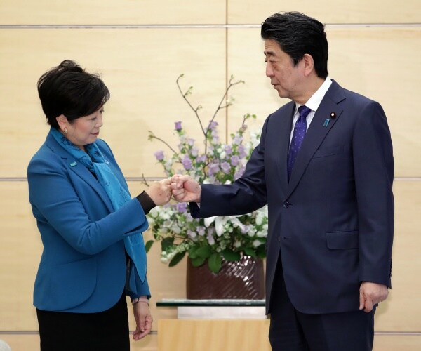 abe in a dark gray suit and blue tie fist bumps koike in a blue coat and scarf