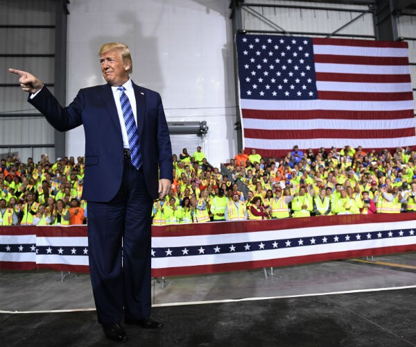 trump smiles and points from stage while a crowd of people wearing yellow protective vests are in the background
