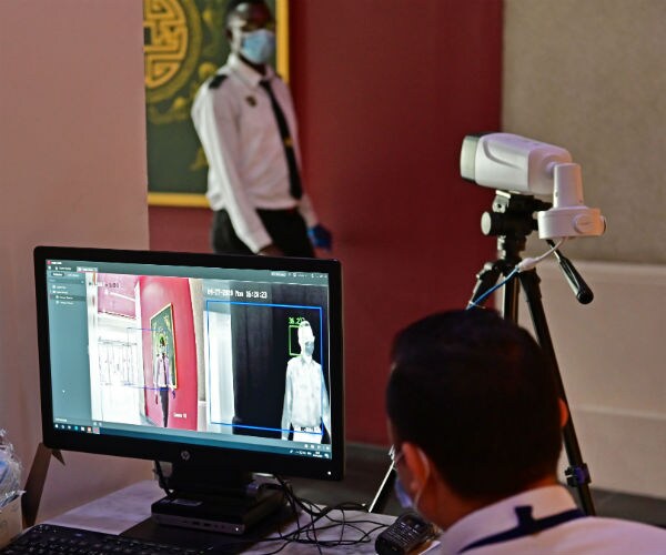 a security guard, wearing a protective mask walks in front of a thermal camera