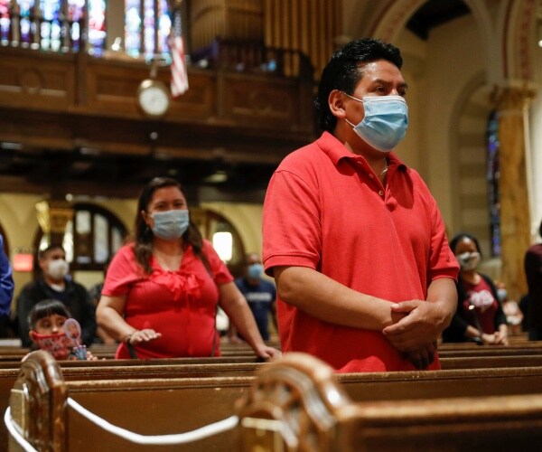 man wears a red shirt and face mask and stands during mass with a woman and child behind him