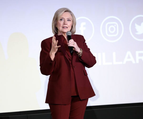 hillary clinton is shown speaking, holding a microphone in a burgundy pantsuit