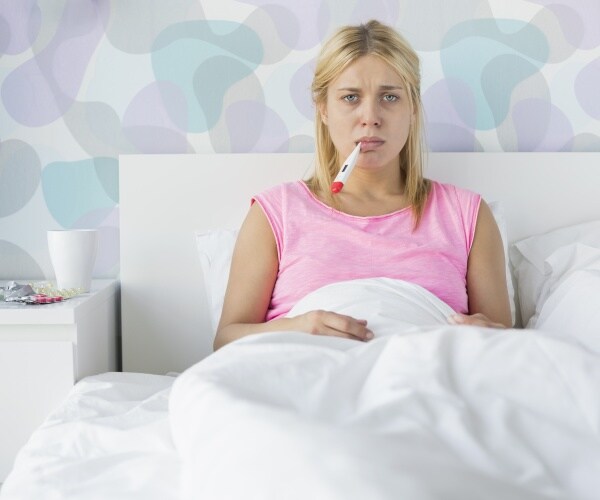woman sitting up in bed looking sick with thermometer in her mouth